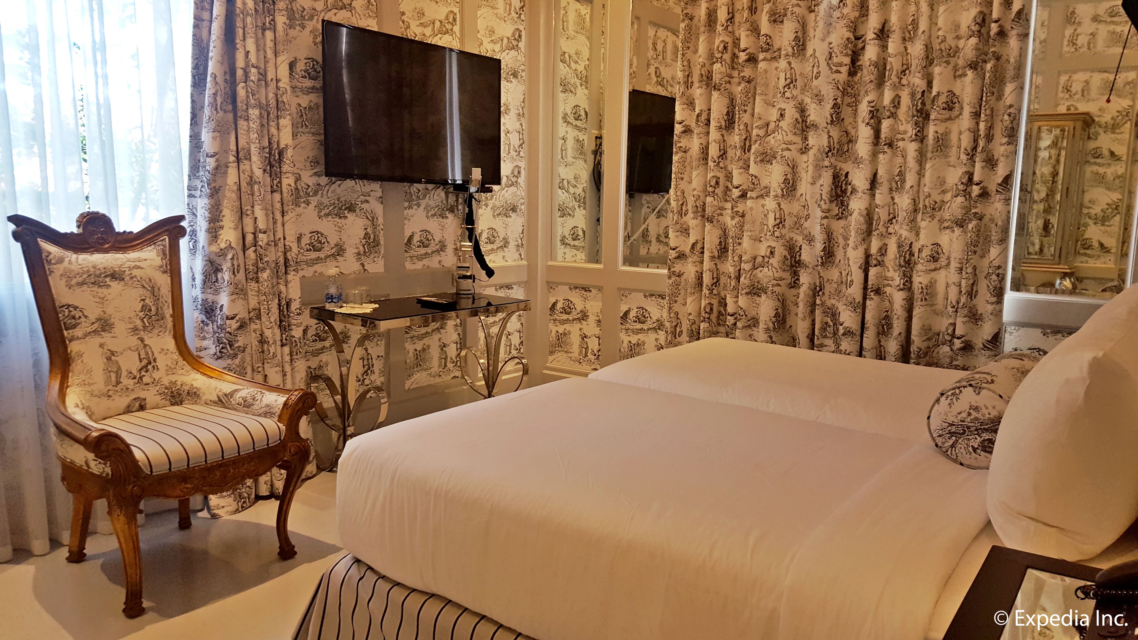 A bedroom with a large bed, a wooden chair, a small table, and a flat-screen TV mounted on the wall.