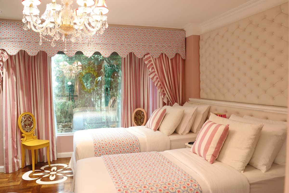A bedroom with two beds, a chandelier, a window with curtains, and a view of greenery outside.