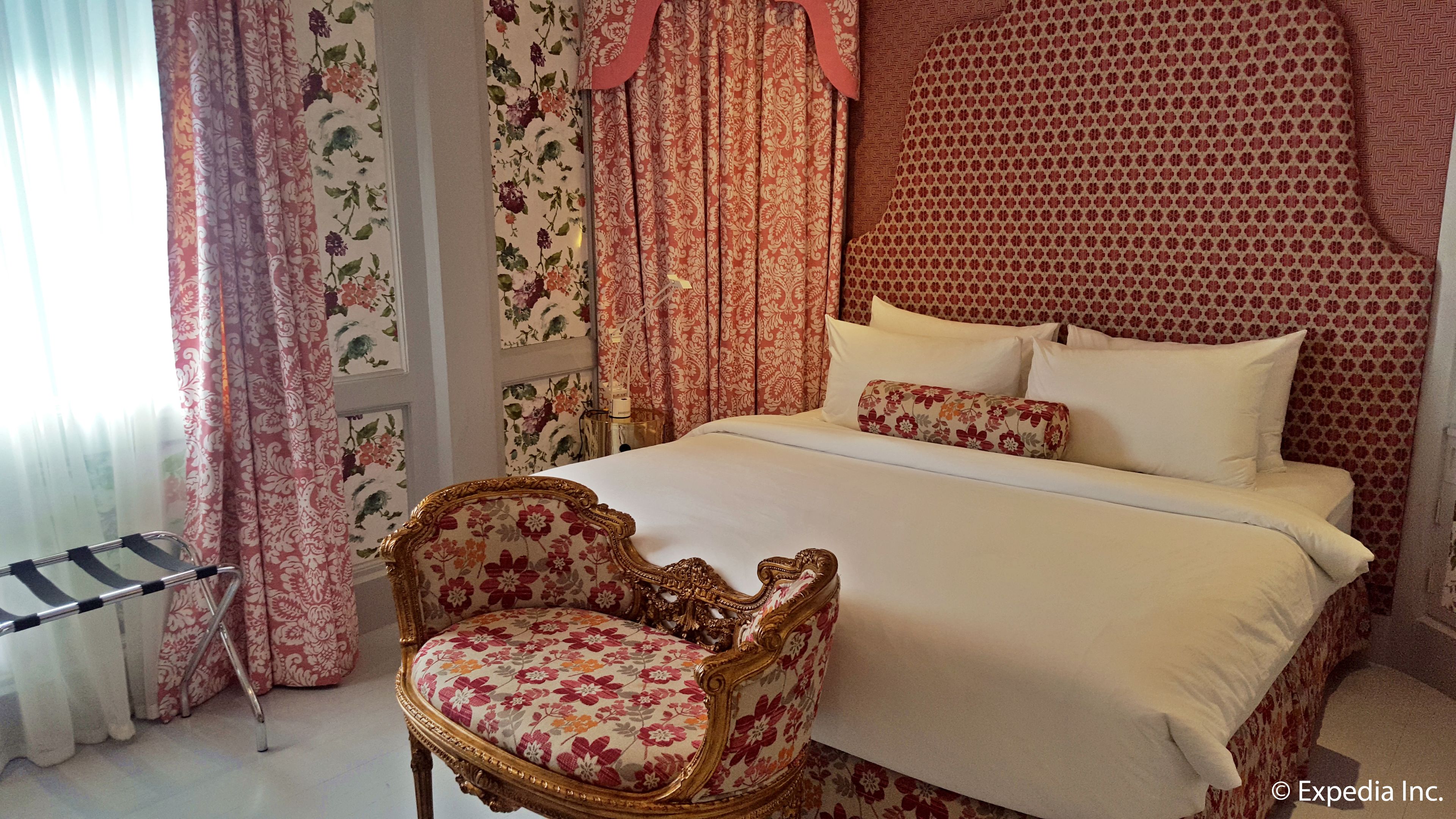 A bedroom with a large bed, floral curtains, a gold-framed chair, and a window with sheer curtains.