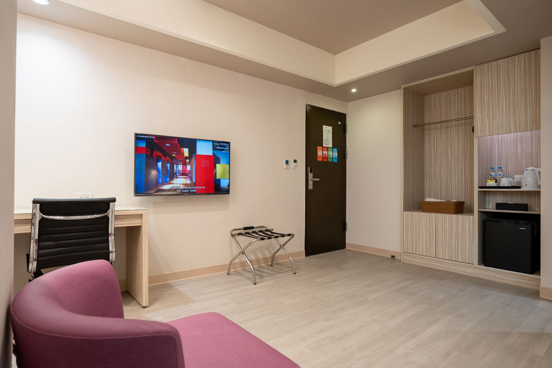 Une chambre d’hôtel équipée d’un téléviseur fixé au mur, d’un bureau avec une chaise, d’un petit coin salon, d’une porte avec une pancarte et d’une kitchenette.