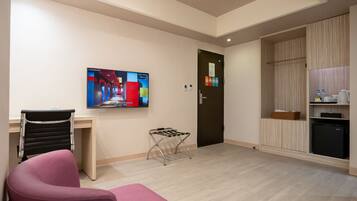 A hotel room with a TV mounted on the wall, a desk with a chair, a small seating area, a door with a sign, and a kitchenette.