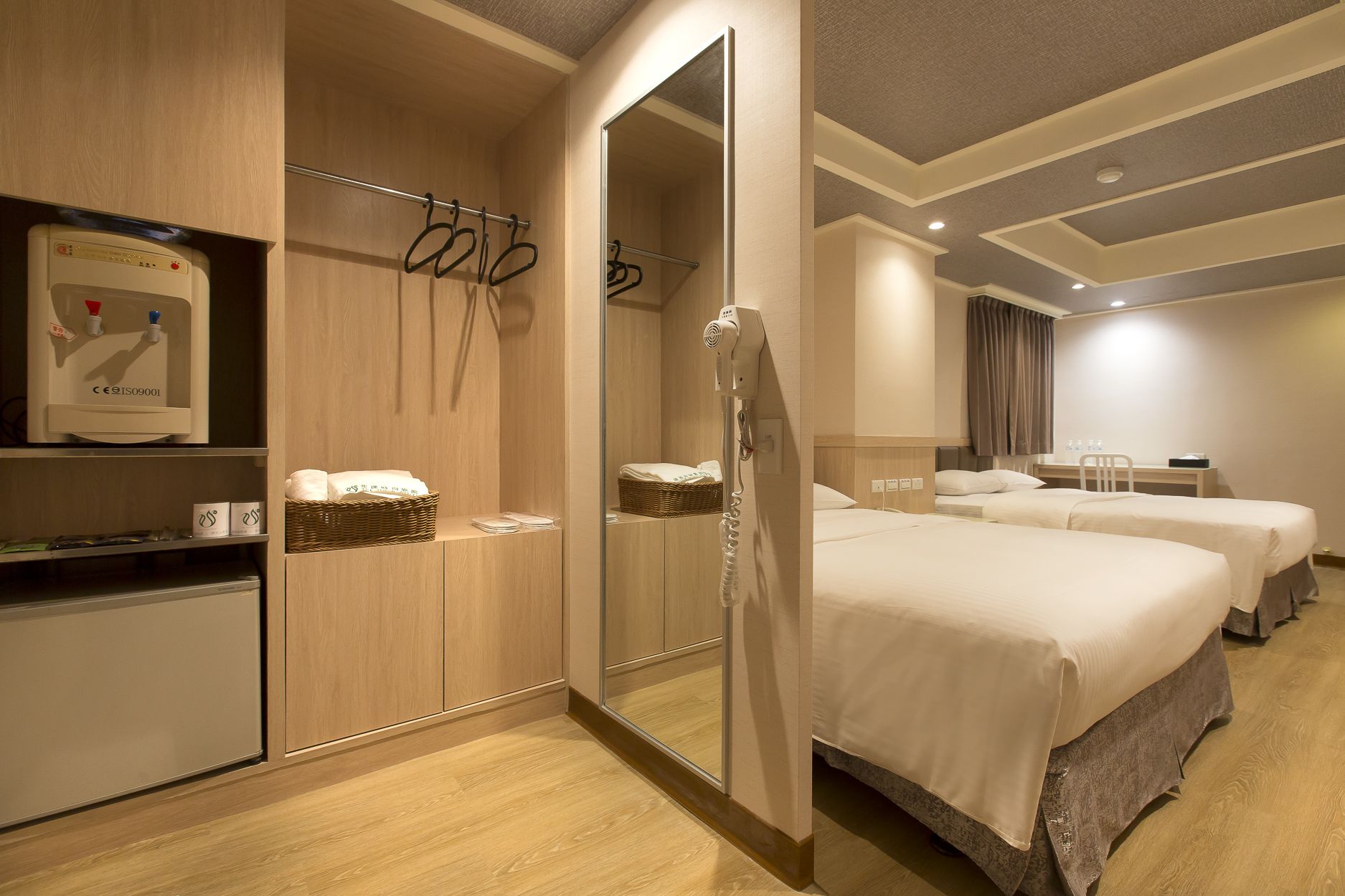 Ein Hotelzimmer mit einem Spiegelkleiderschrank, einem Fön, einer Minikühlschrank, einem Nachttisch mit Korb und Handtüchern sowie zwei Betten.