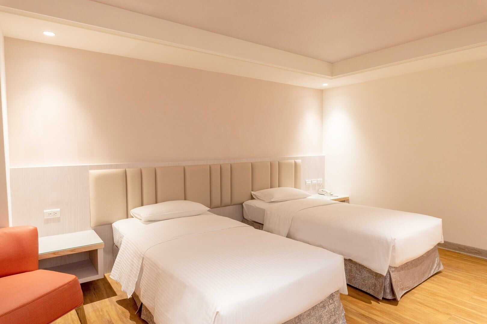 Habitació d'hotel amb dos llits, una tauleta de nit, una cadira vermella i un terra de fusta.