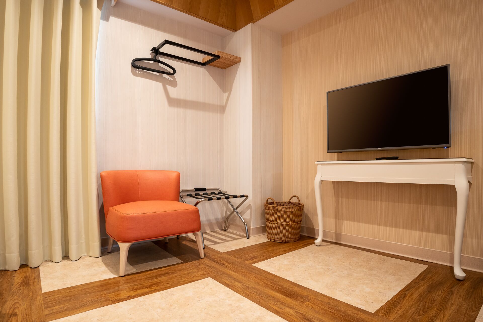 A modern living room with a flat-screen TV on a white console table, a single orange chair, a small side table, and a wicker basket.
