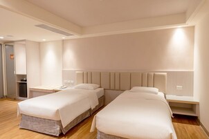 Una habitación de hotel con dos camas individuales, una mesita de noche con lámpara, un escritorio pequeño y un armario.
