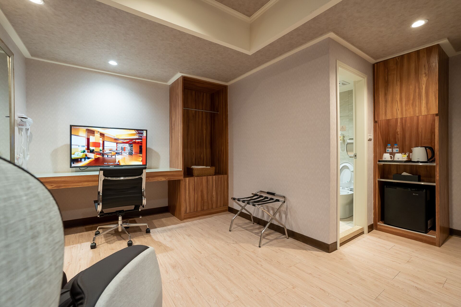 A hotel room with a desk, chair, TV, wooden wardrobe, and a small kitchenette.