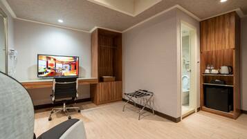 A hotel room with a desk, chair, TV, wooden wardrobe, and a small kitchenette.