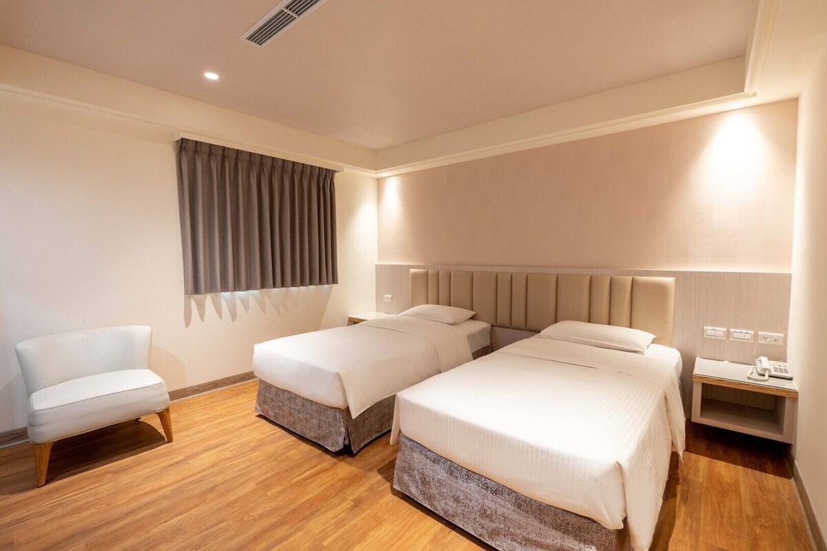 Une chambre d’hôtel avec deux lits, une chaise, une table de chevet avec une lampe et une fenêtre avec des rideaux.