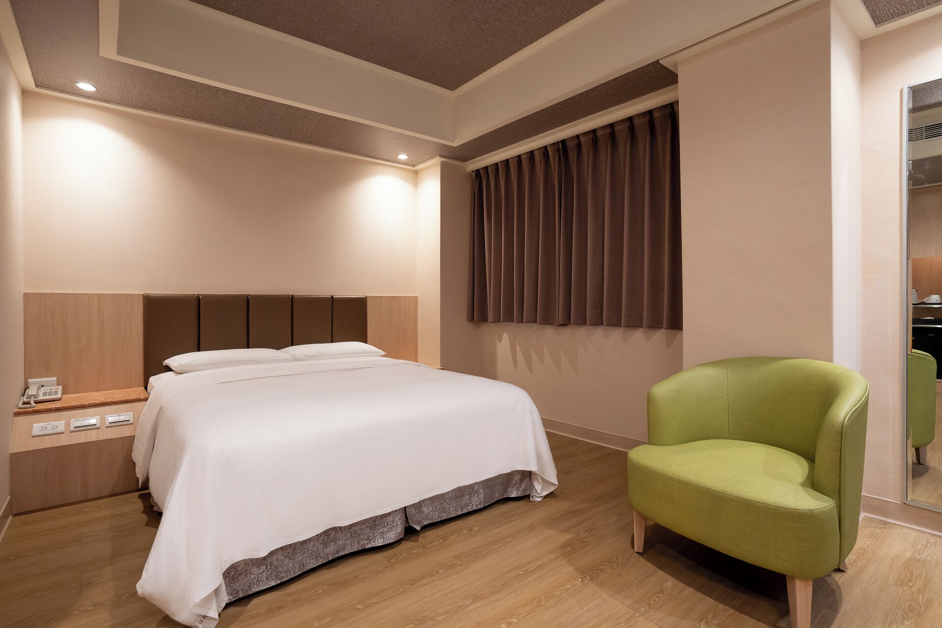A hotel room with a bed, a green armchair, a bedside table with a phone, and a window with curtains.