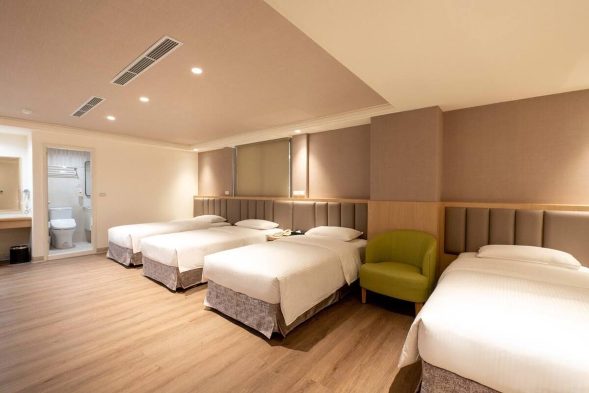 Une chambre d’hôtel avec quatre lits, un fauteuil vert et une salle de bain visible à travers une porte ouverte.