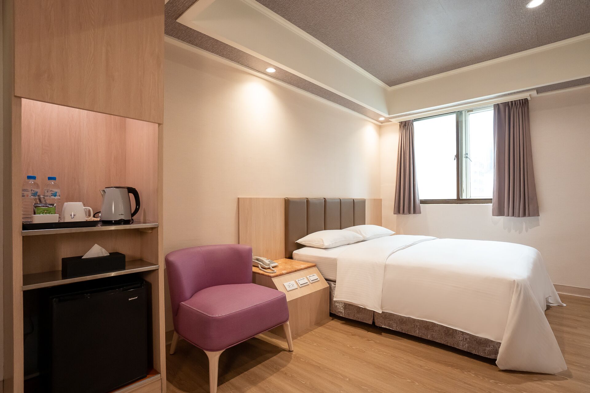 A hotel room with a bed, a desk with a chair, a mini-fridge, a coffee maker, and a window with curtains.