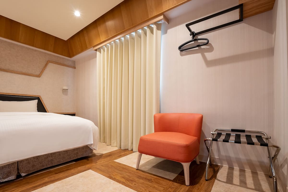 Une chambre d’hôtel avec un lit, un fauteuil orange, un étagère en métal et une fenêtre avec des stores verticaux.