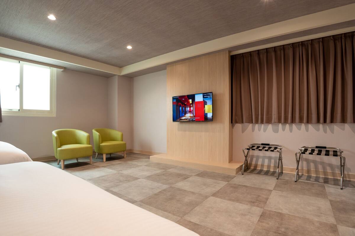 Une chambre d’hôtel avec un grand lit, deux fauteuils verts, un téléviseur fixé au mur et une fenêtre avec des rideaux.