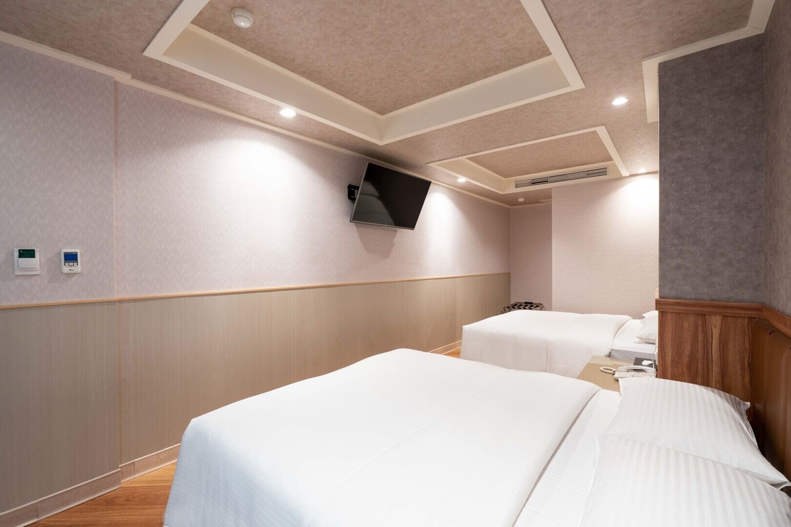A hotel room with two beds, a flat-screen TV mounted on the wall, and a ceiling with recessed lighting.