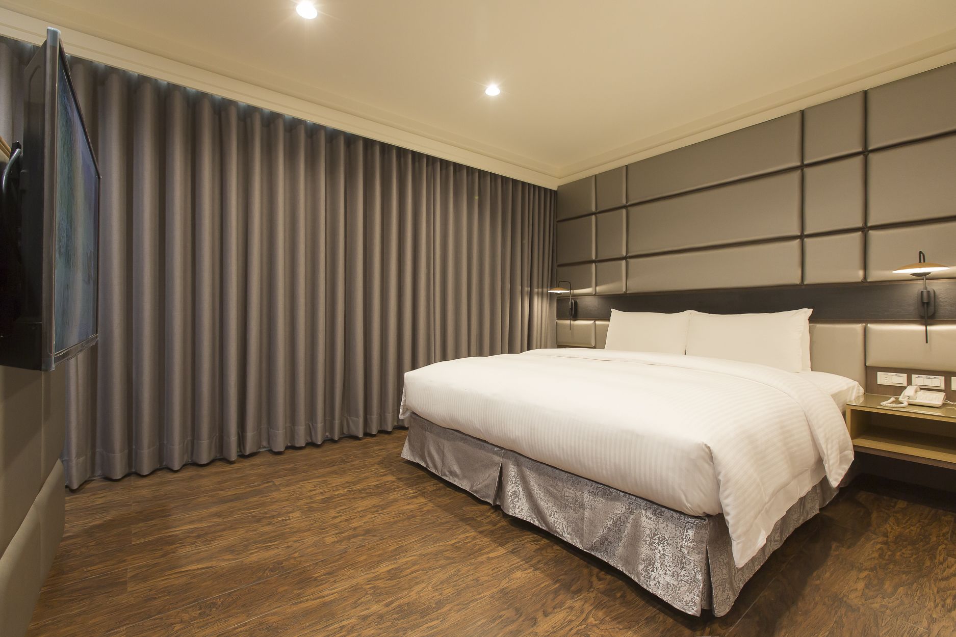 Una moderna camera d'albergo con un letto grande, una TV a schermo piatto fissata al muro e un comodino con lampada e telefono.