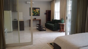 A hotel room with a bed, a green sofa, a small table, a painting on the wall, and a kitchen area visible through a glass door. - Kemang Inn (Jakarta)