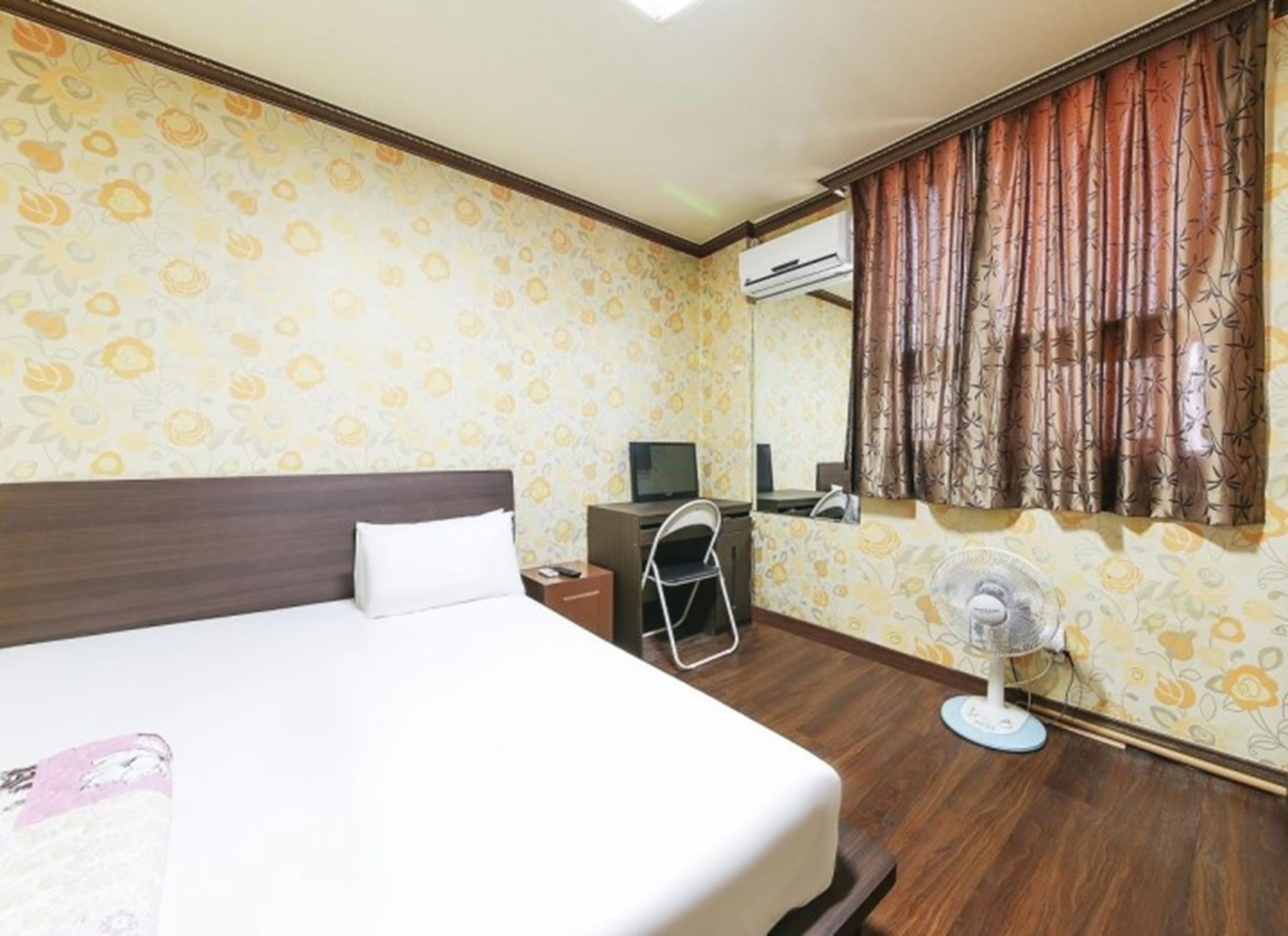 Una camera d'albergo con un letto, una scrivania, una sedia e una ventola da pavimento.