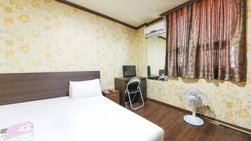 Una camera d'albergo con un letto, una scrivania, una sedia e una ventola da pavimento.