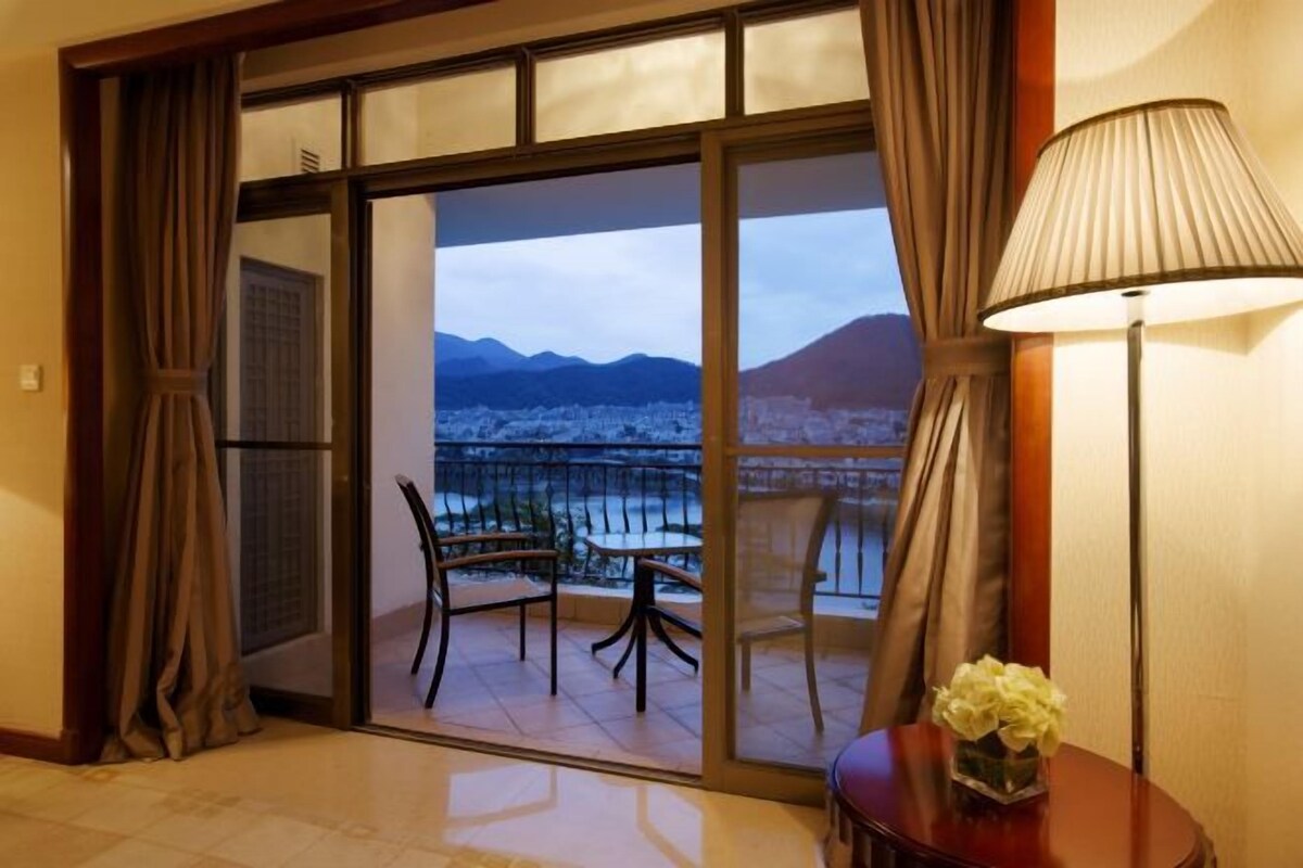Una camera d'hotel con un balcone, un tavolo con sedie, una lampada e un vaso di fiori.
