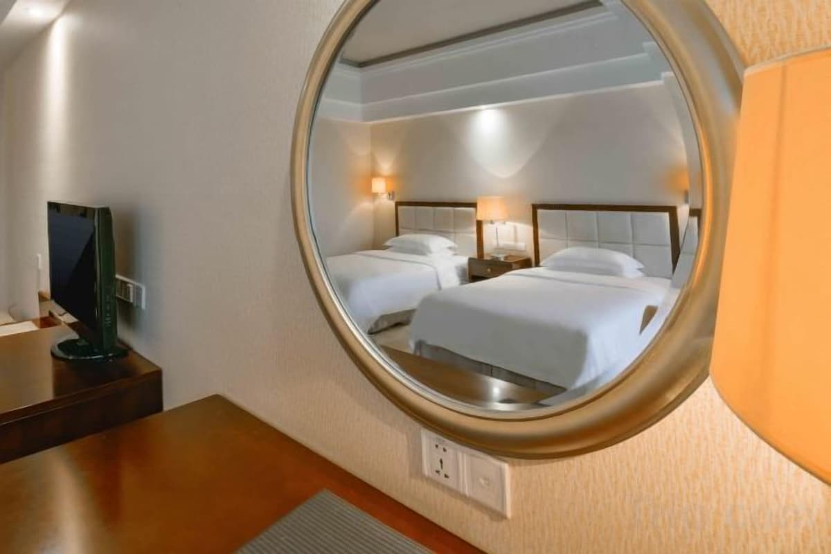 A hotel room with a round mirror reflecting two beds, a bedside table, and a television.