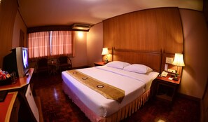 A hotel room with a bed, a desk with a television, two chairs, a nightstand with a lamp, and a wooden headboard. - Silom Village Inn (Bangkok)