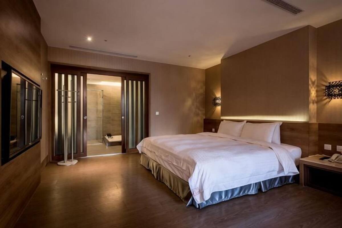 Une chambre d’hôtel avec un grand lit, une table de chevet, une commode, une lampe fixée au mur et une porte coulissante donnant accès à la salle de bain.