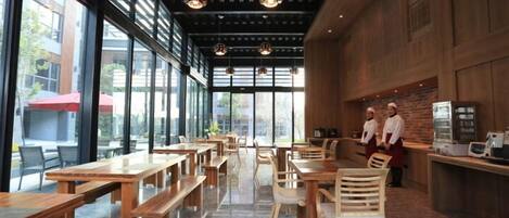 Un comedor interior con mesas y sillas de madera, ventanas amplias y una pared de ladrillo.