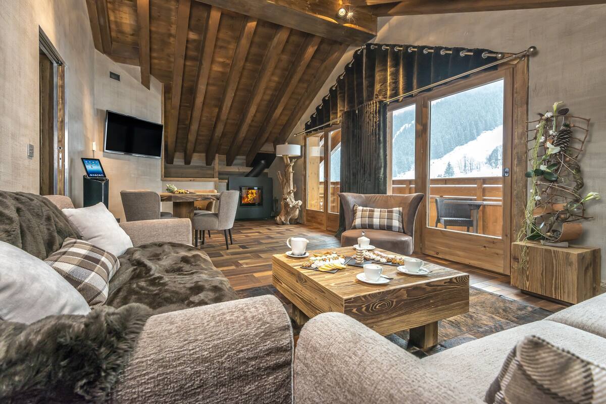 Una sala de estar acogedora con techo de madera, chimenea, comedor con mesita de centro y vistas a montañas nevadas a través de amplias ventanas.