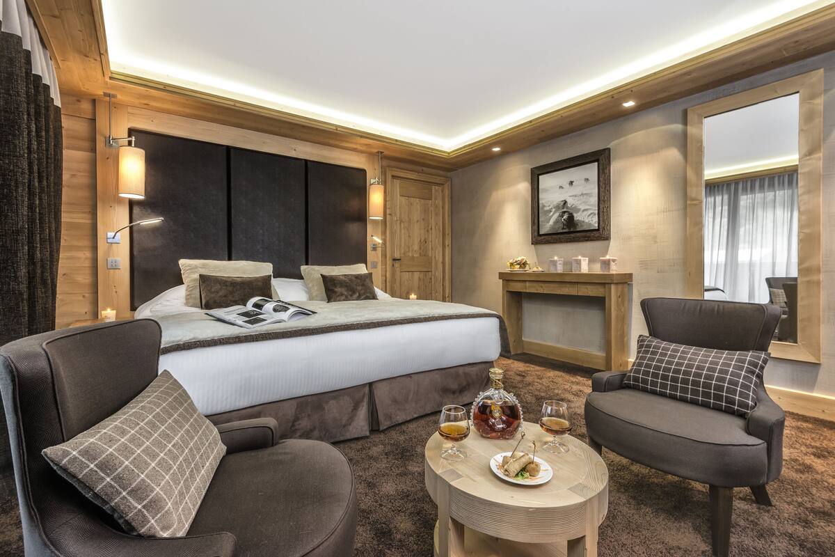 Una habitación de hotel moderna con una cama grande, dos sillones, una mesita redonda con bebidas y aperitivos, y una chimenea.