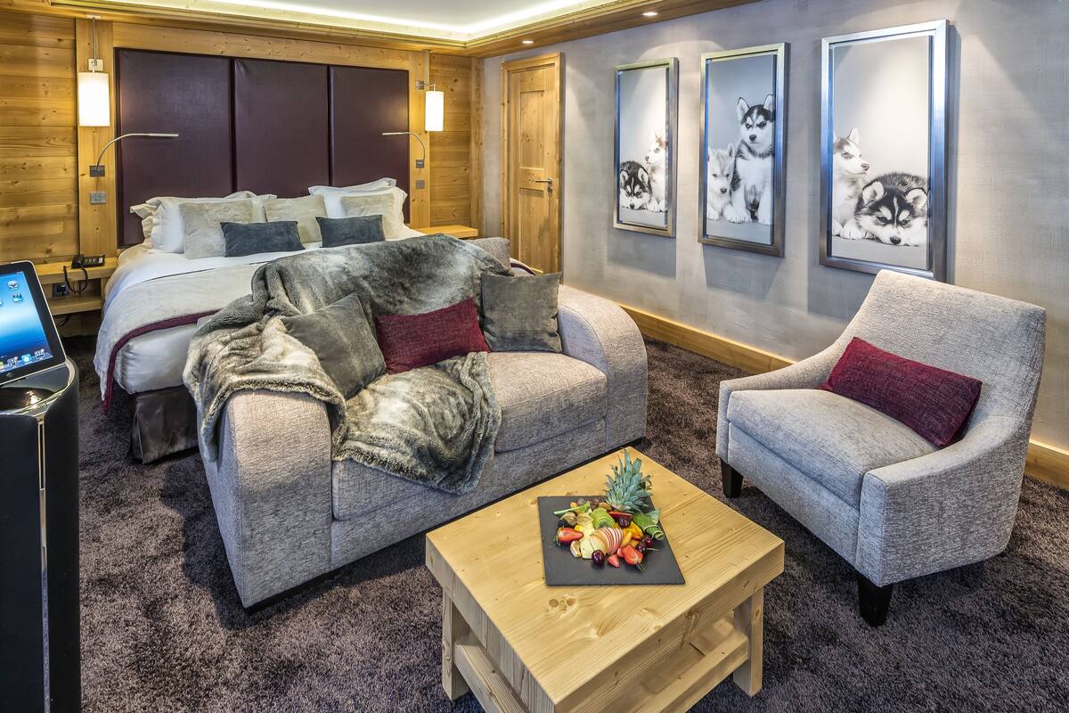 Habitación de hotel con una cama, un sofá gris, una mesa de centro de madera con fruta y cuadros en la pared.