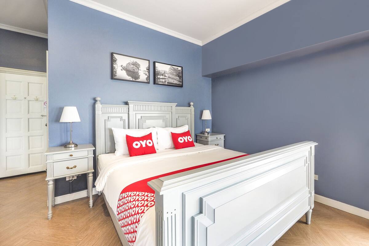 Quarto com cama branca, duas almofadas vermelhas, mesa de cabeceira branca, penteadeira branca e parede azul.