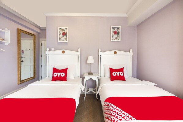 Habitación de hotel con dos camas, cada una con cabecera blanca y almohadas rojas con la palabra 'OYO'. Hay una mesita con una lámpara entre las camas, y un espejo en la pared izquierda.