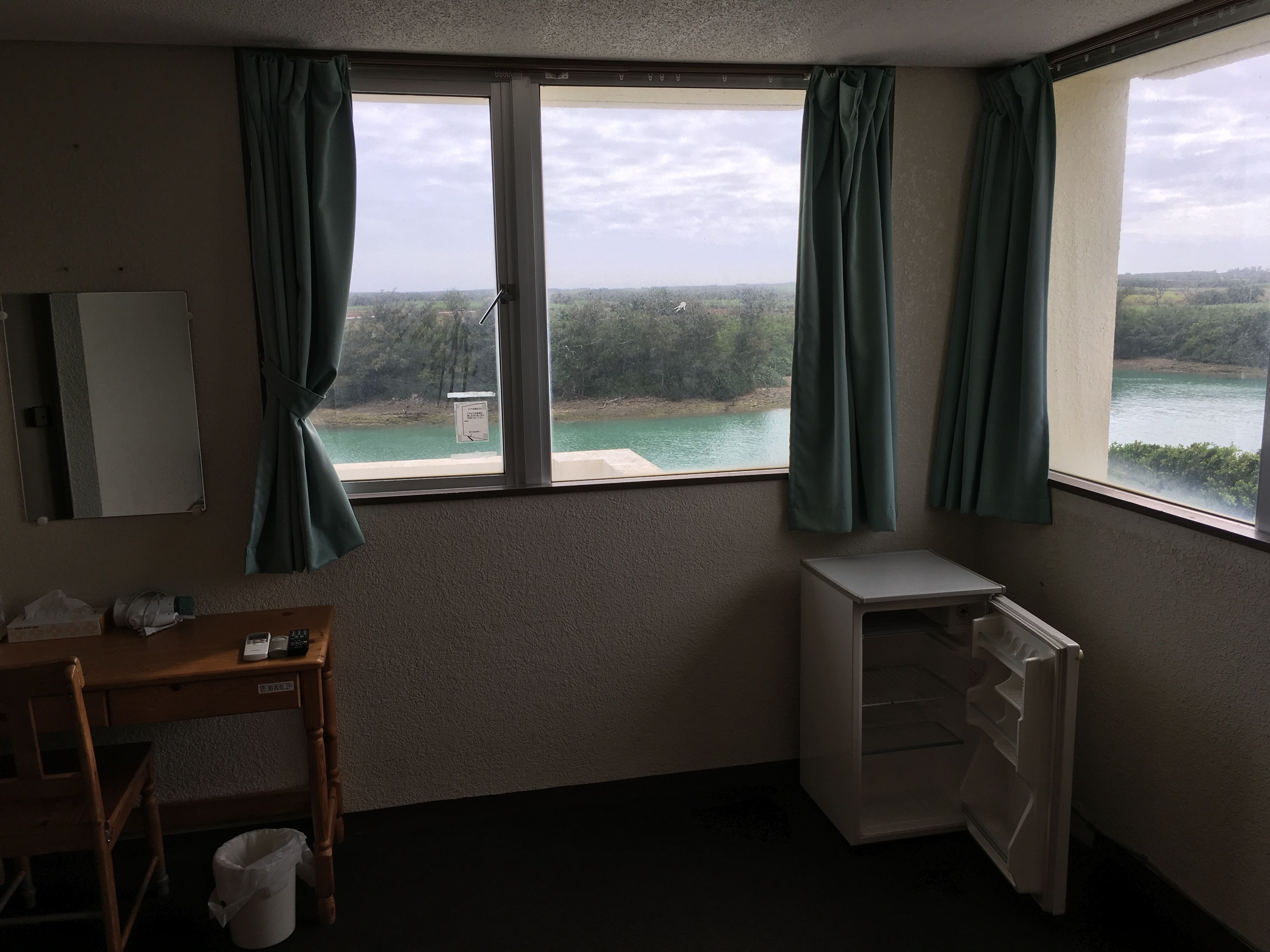 Hotelkamer met uitzicht op een meer, een houten bureau, een stoel, een klein wit kastje en een prullenbak.