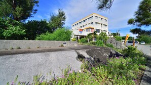 Un edificio de varios pisos con fachada blanca y detalles anaranjados, rodeado de vegetación y una barrera de concreto.