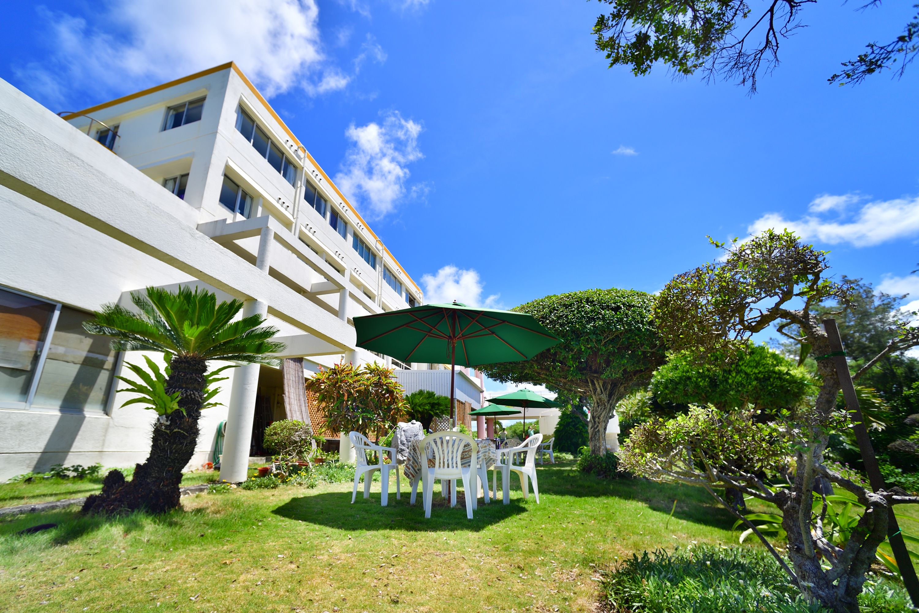 A modern building with a white facade and a well-maintained garden with a table and chairs under a green umbrella.