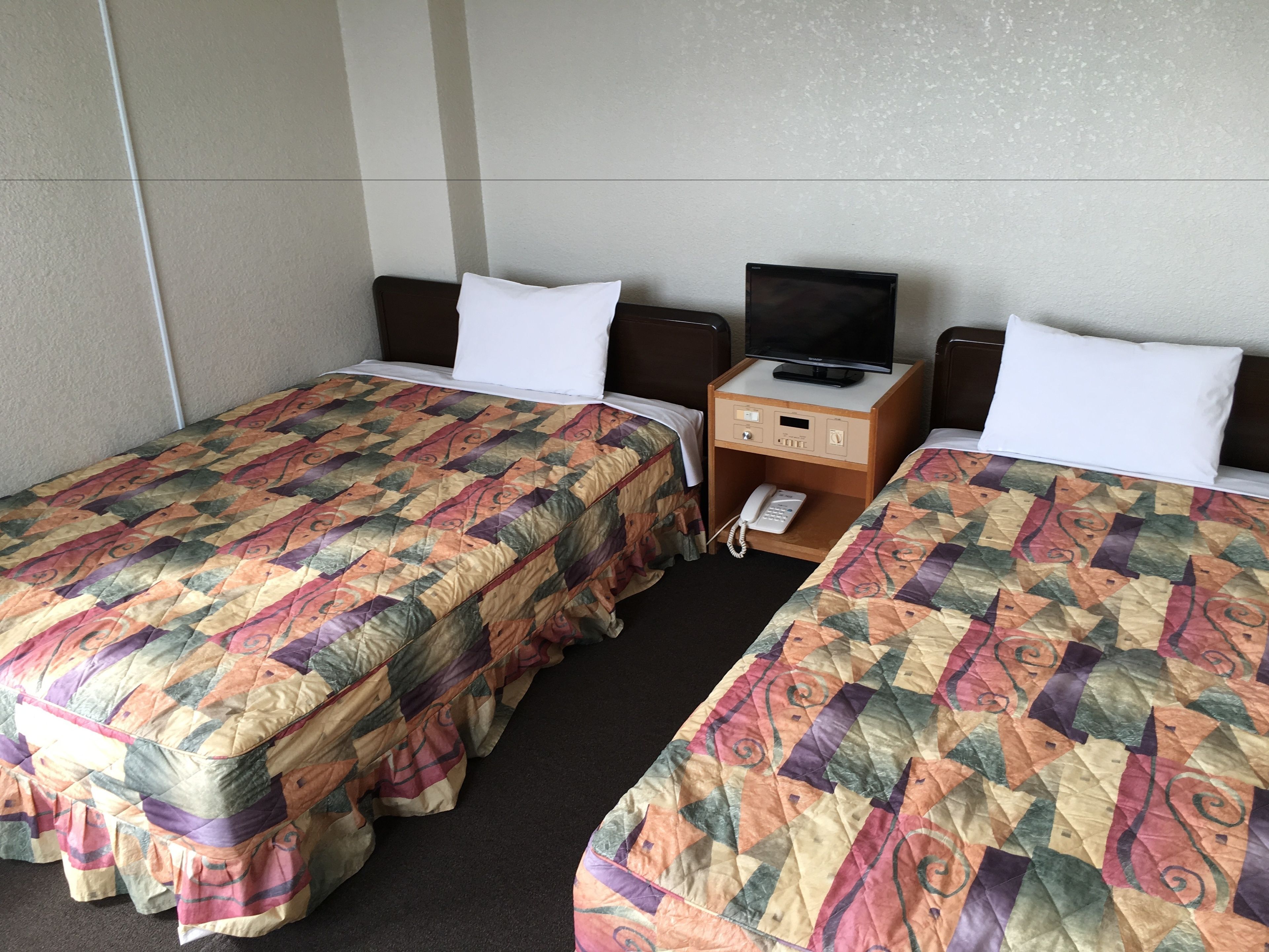 A hotel room with two beds, each with a colorful quilt, a small wooden nightstand with a television and a phone, and white pillows.