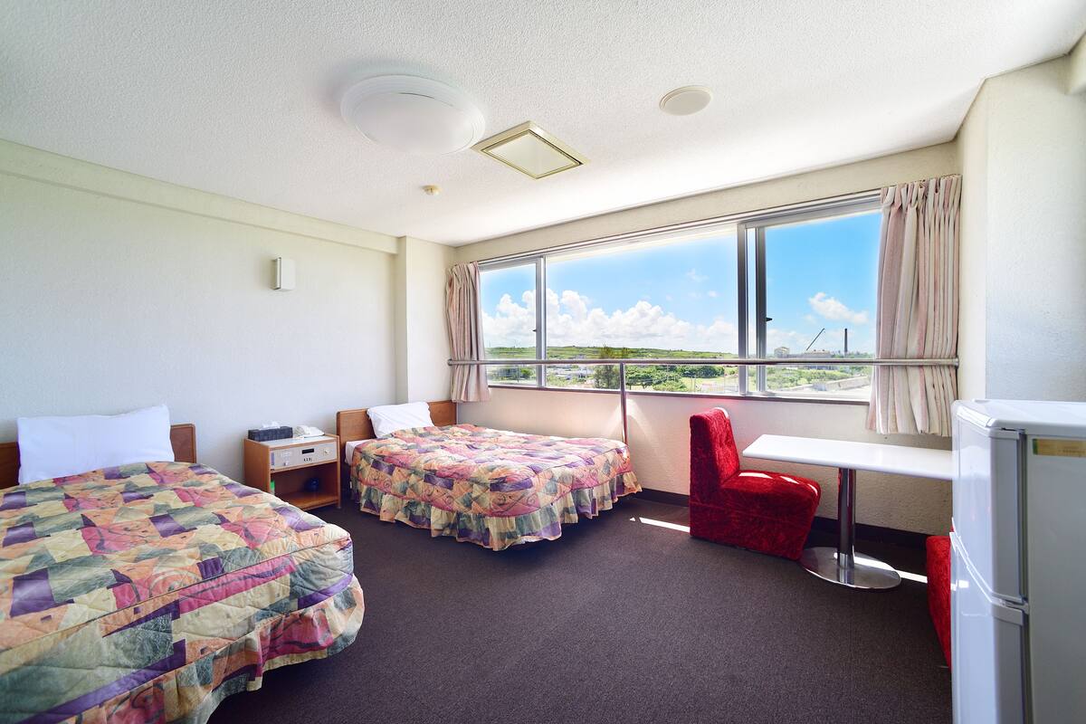 A hotel room with two beds, a desk, a chair, a refrigerator, and a large window with a view.