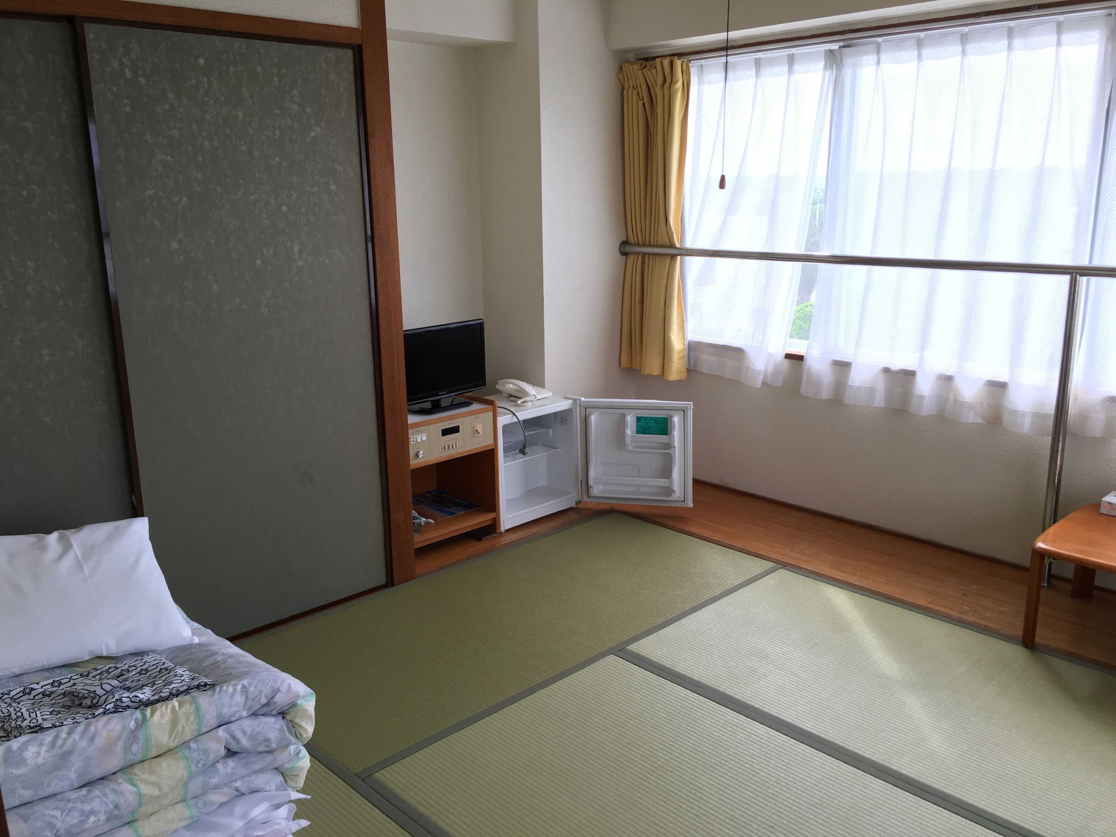 A hotel room with a bed, a TV, a mini-fridge, a bedside table, and a window with curtains.