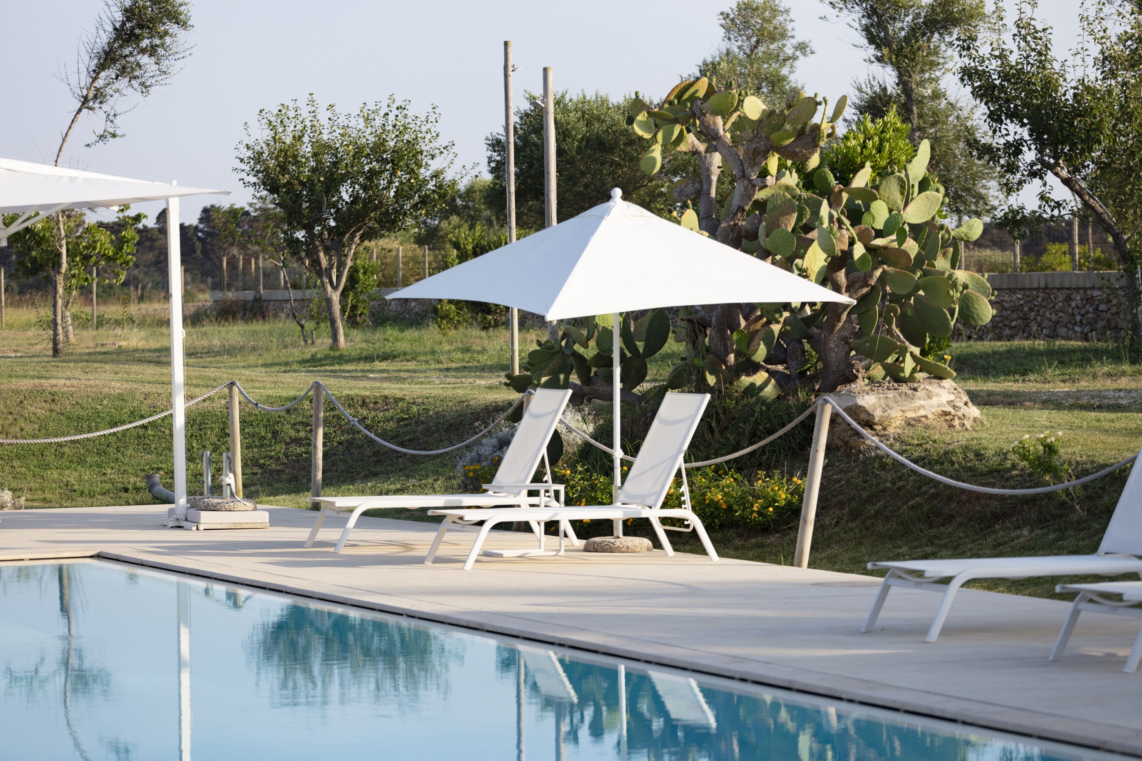 A poolside area with lounge chairs and umbrellas, surrounded by greenery and a stone wall.