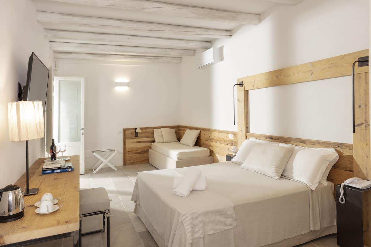 Une chambre d’hôtel avec une tête de lit en bois, un lit avec du linge de maison blanc, une table de chevet avec une lampe, une petite table avec une bouilloire et une chaise.