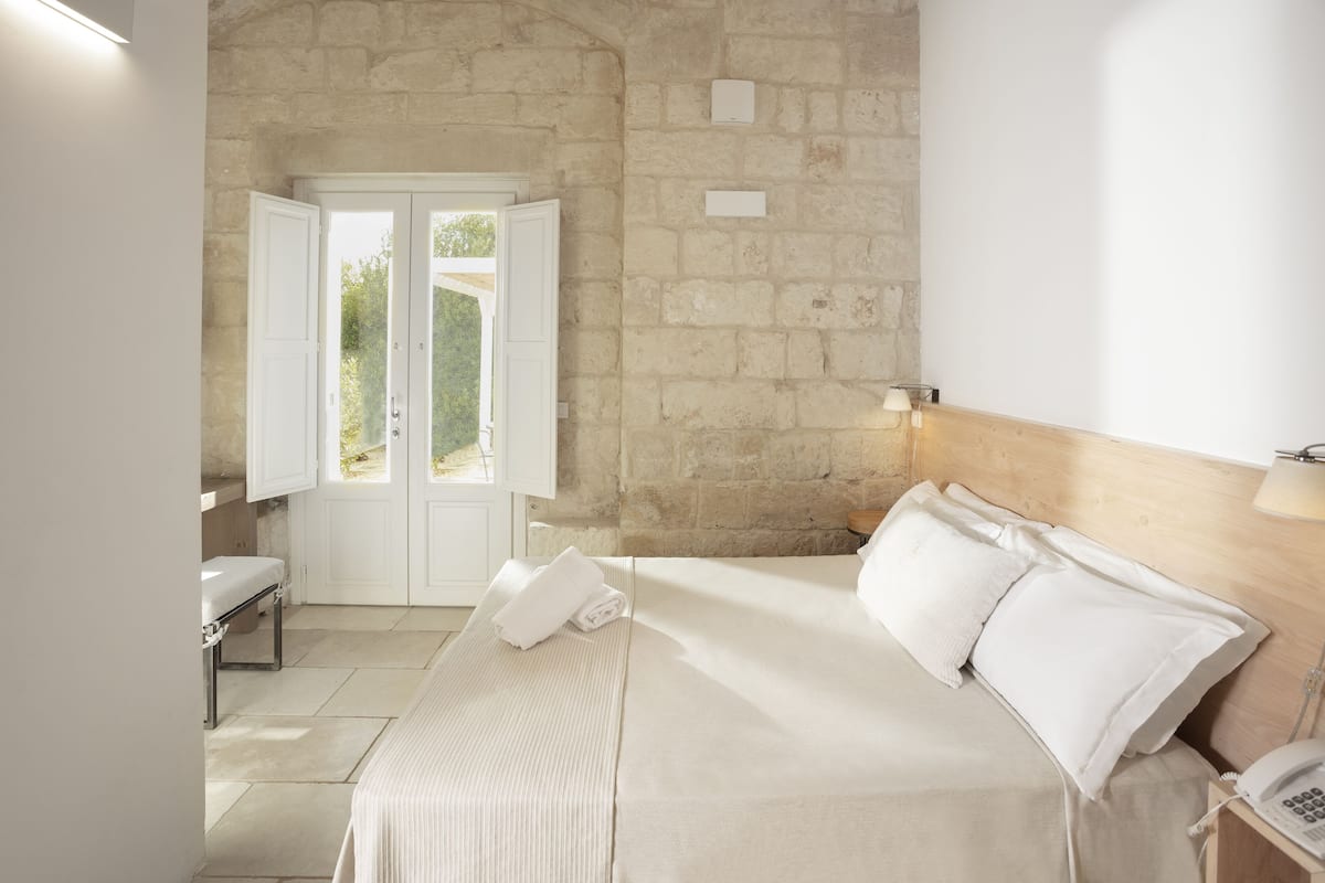 Une chambre avec un mur en pierre, un lit recouvert de draps blancs, une table de chevet avec une lampe et une fenêtre dont les volets sont ouverts.