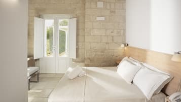 Un dormitorio con pared de piedra, cama con ropa blanca, mesita de noche con lámpara y ventana con persianas abiertas.
