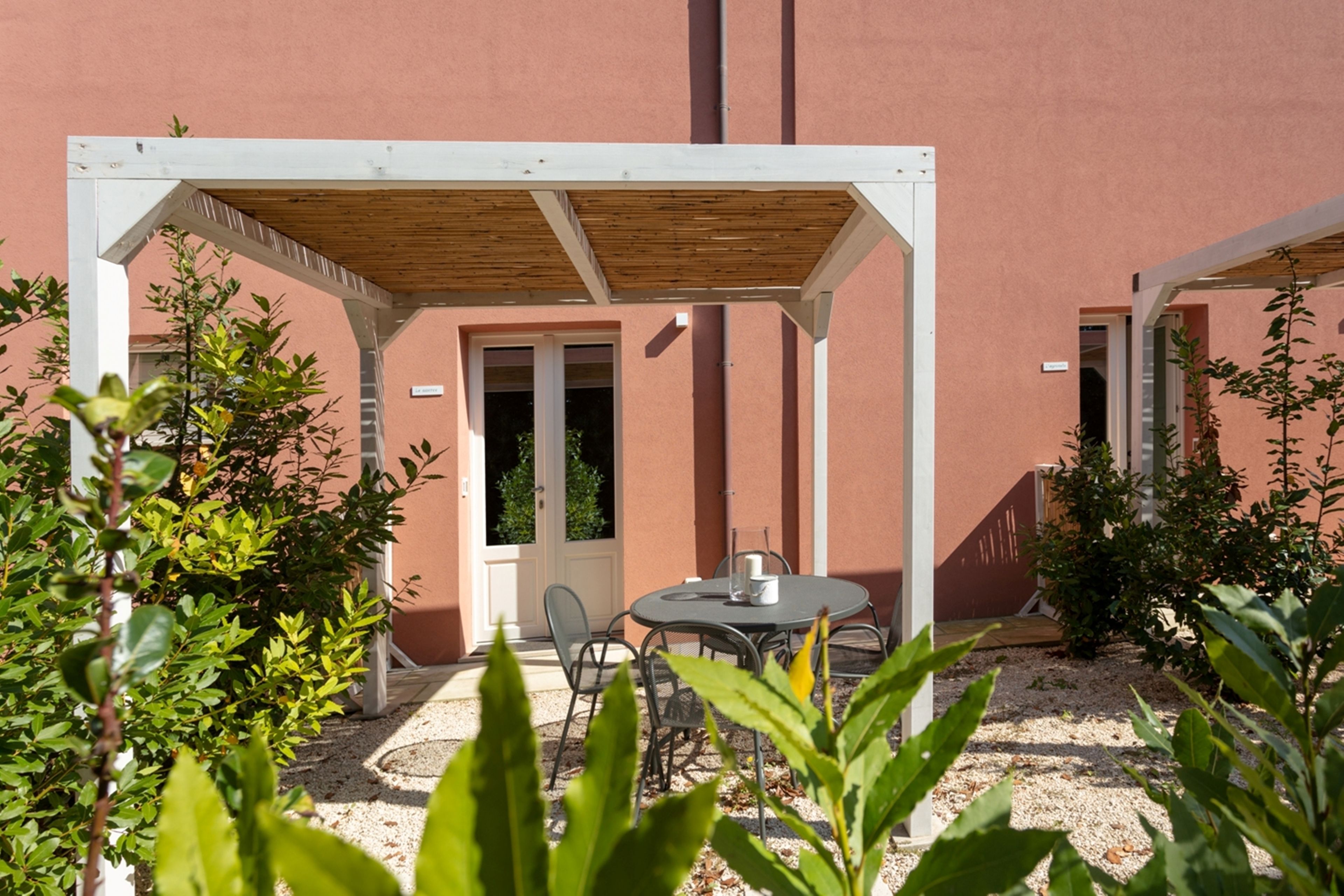 A covered outdoor seating area with a table and chairs, surrounded by plants and a building with a red exterior.