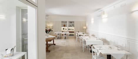 A dining area with white tables and chairs, a long counter, and a display of wine bottles in the background.