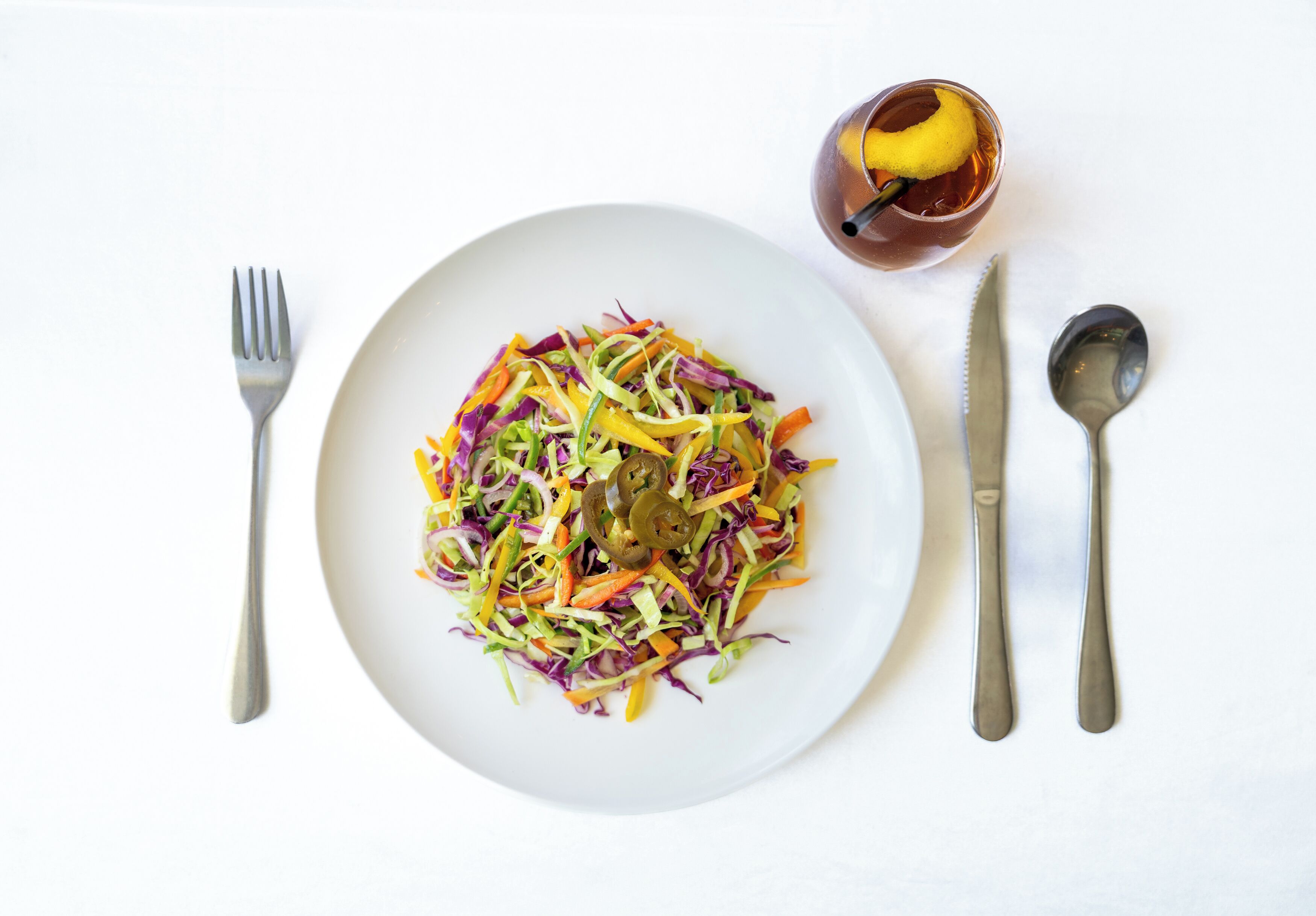 A white plate with a colorful salad, a glass with a lemon slice, and cutlery on a white surface.