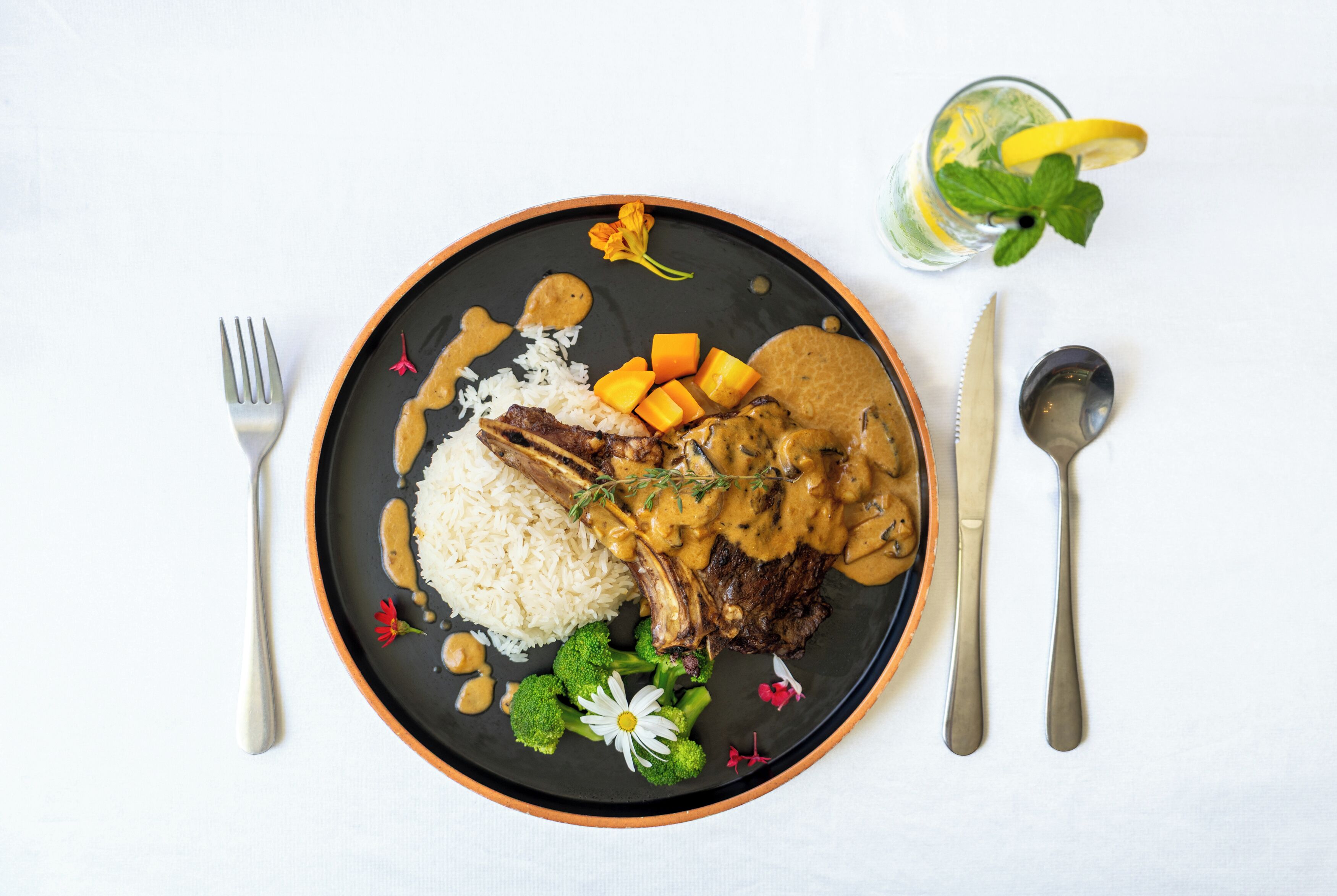 A plate with rice, meat, and vegetables, accompanied by cutlery and a glass of drink.