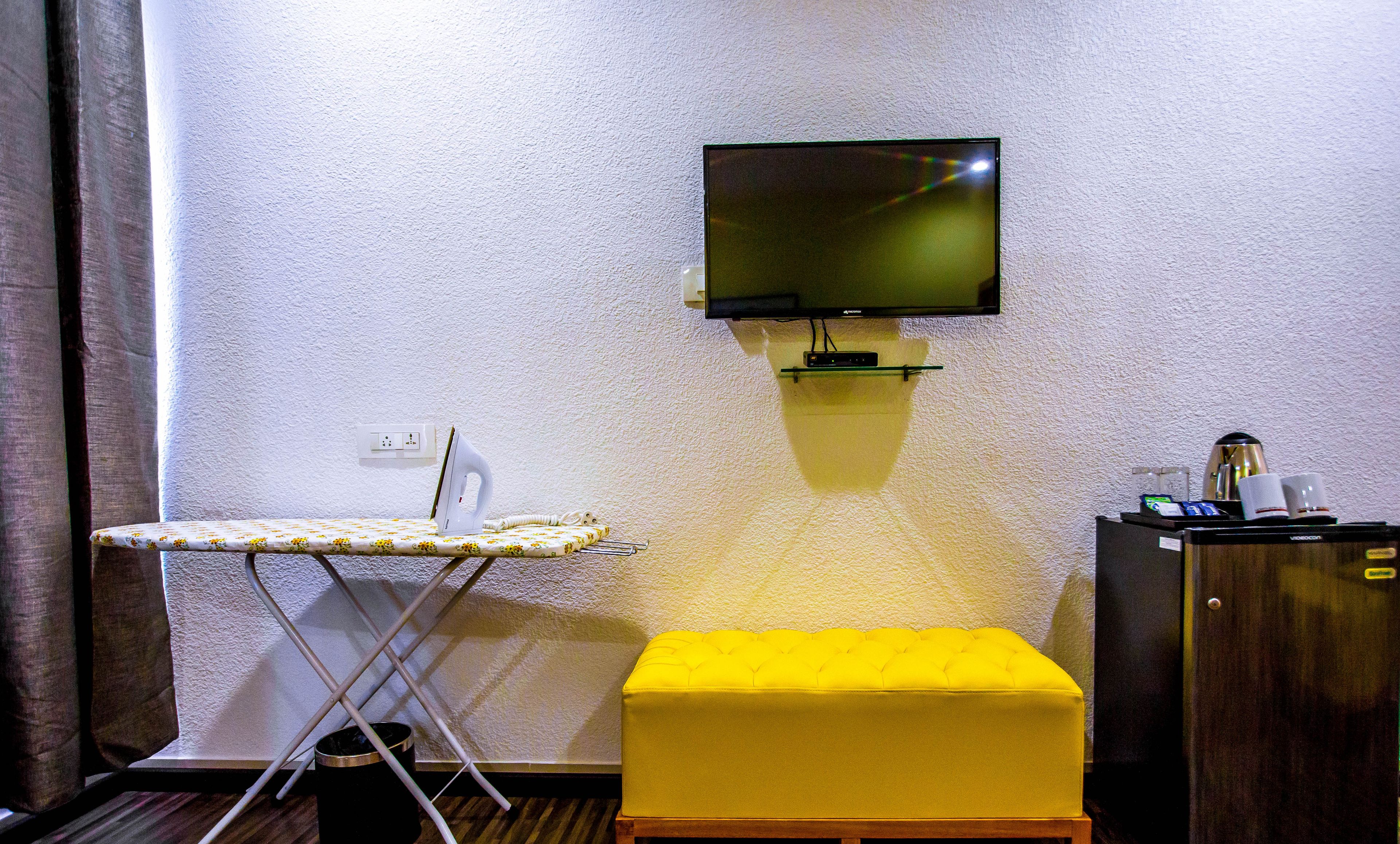 A small room with a television mounted on the wall, a small ironing board, a yellow ottoman, and a cabinet with a coffee maker.