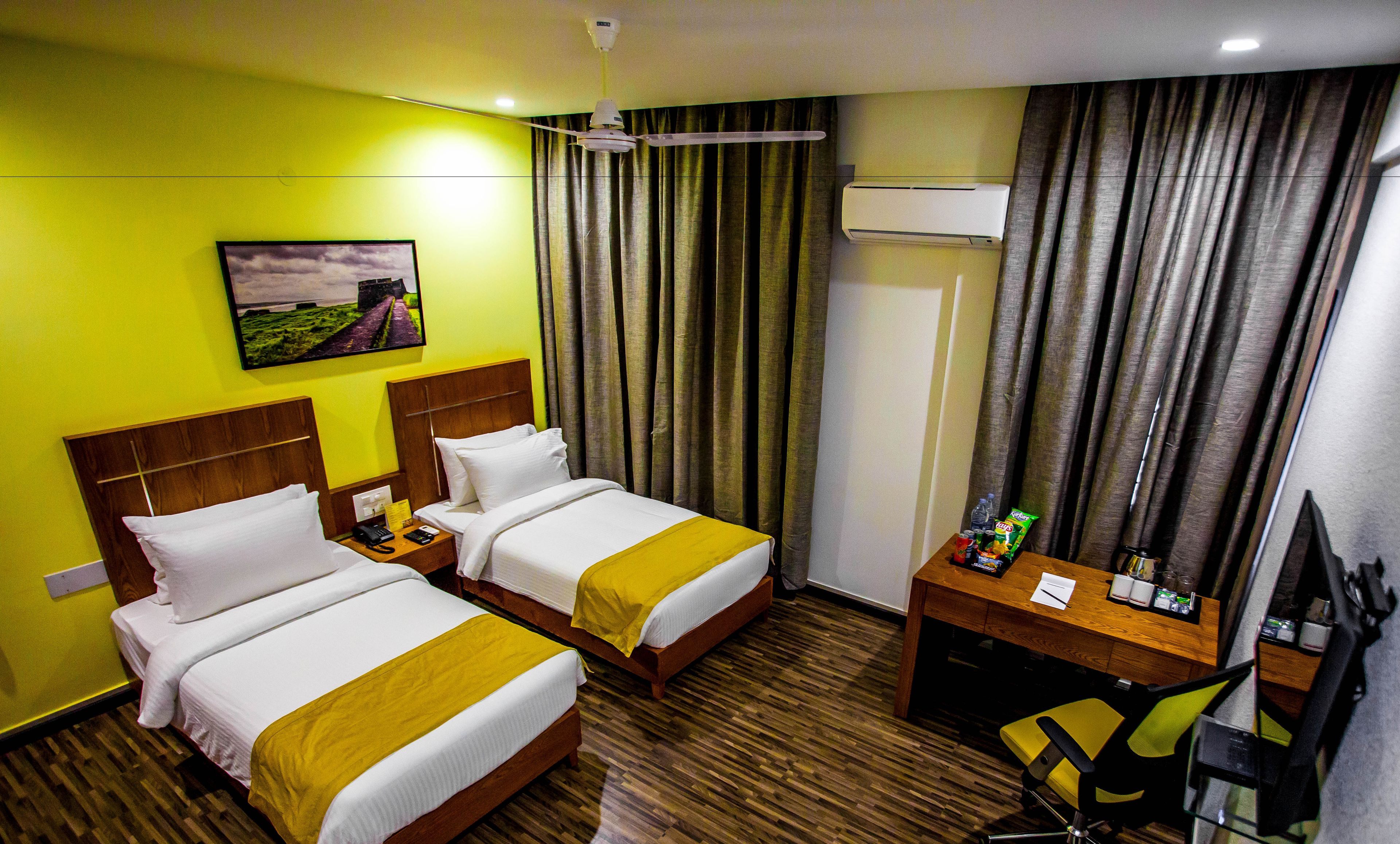 A hotel room with two beds, a desk, a chair, an air conditioner, and a painting on the wall.