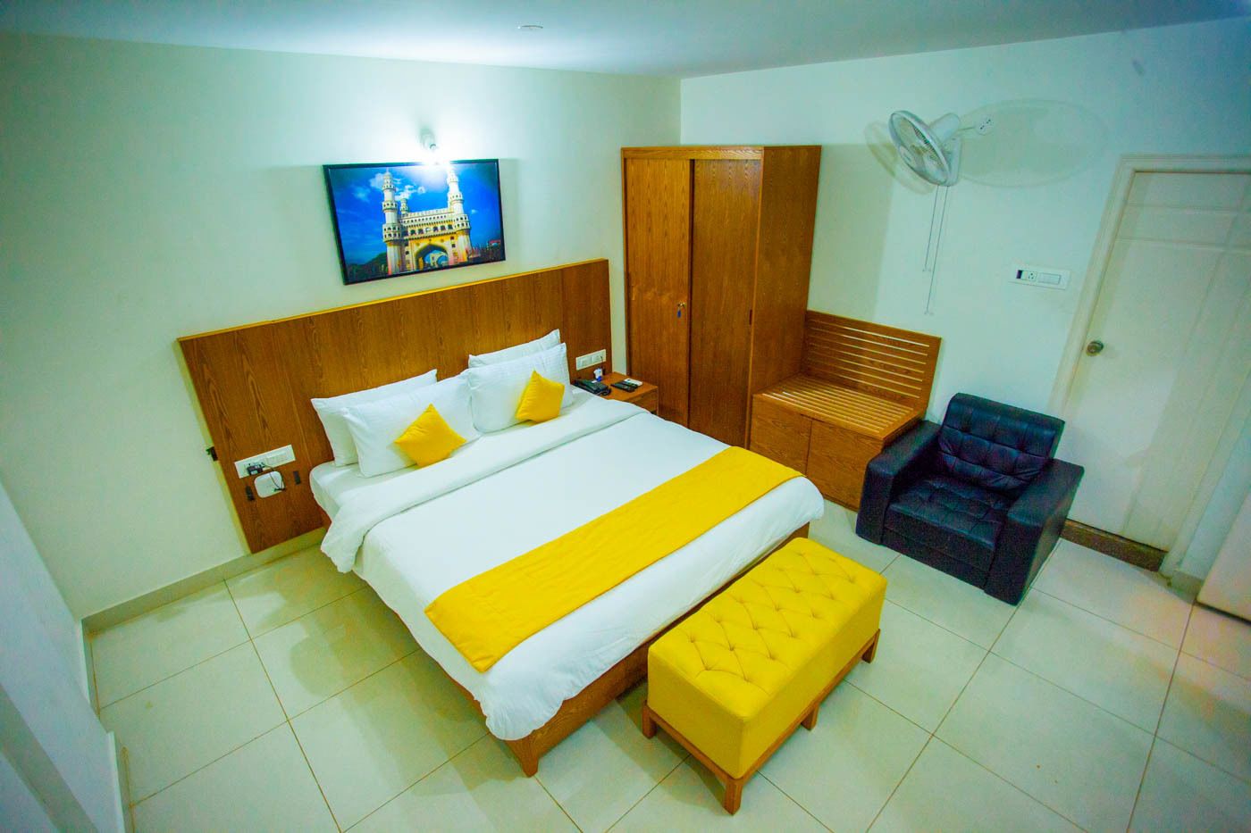 Een hotelkamer met een bed, een houten garderobe, een bankje, een stoel en een ventilator aan de muur.