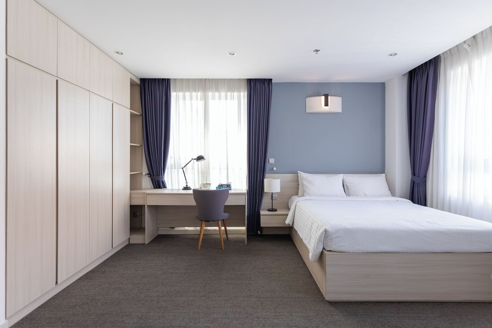 A modern hotel room with a bed, desk, chair, and wardrobe.